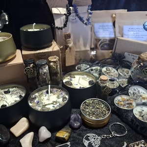 May include: A collection of black and gold metal candle tins filled with wax, dried herbs, and crystals. Includes small glass vials with cork stoppers, tea light candles, a tree of life keychain, and various loose crystals arranged on a dark surface.