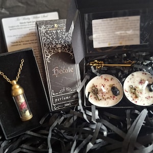 May include: A gift set featuring a gold-toned vial necklace, a 30ml bottle of Hecate perfume oil, and a black box containing two white tea light candles topped with dried flower petals, small metal keys, and black stones, plus a gold skeleton key.