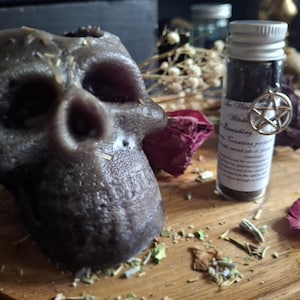 May include: A dark gray skull-shaped candle with a textured surface, positioned next to a small clear glass vial containing dark powder. The vial features a white label with black text and a silver pentagram charm attached to the neck.