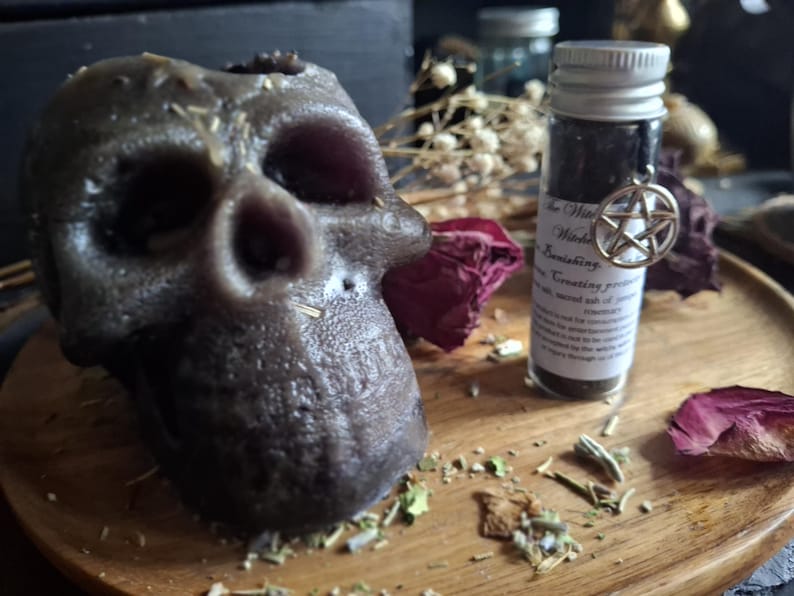 May include: A dark gray skull-shaped candle with a textured surface, positioned next to a small clear glass vial containing dark powder. The vial features a white label with black text and a silver pentagram charm attached to the neck.