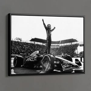 May include: Black and white framed canvas print featuring a Formula 1 race car with a driver standing on top, arms raised in victory. The image is set against a backdrop of a crowded stadium. The text "BLACK FRAMED CANVAS" is at the bottom.