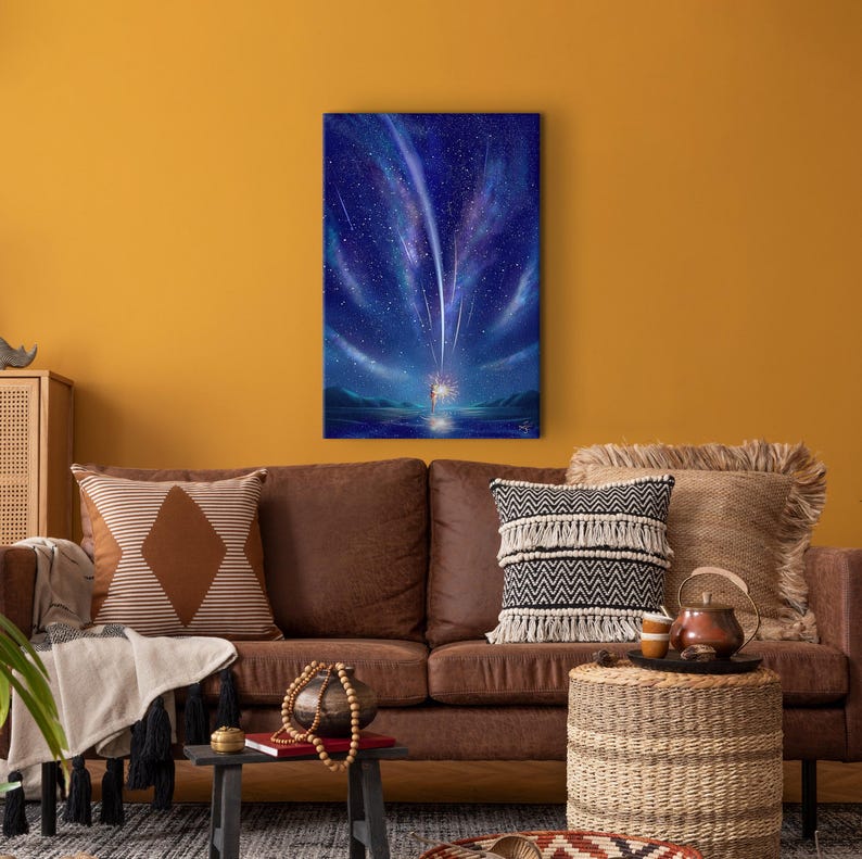 May include: A painting of a starry night sky with shooting stars and a glowing light in the distance. The painting is framed and hanging on a yellow wall above a brown leather couch.