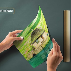May include: A canvas rolled poster featuring a painting of a small house with a thatched roof, surrounded by green grass and water. The house is painted in a light brown color with white trim. The water is a deep blue color and the grass is a bright green. The poster is rolled up and being held by a person's hands.