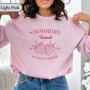 May include: Light pink sweatshirt with the text "STRAWBERRY Festival" and two strawberry illustrations. Below the strawberries, the text reads "LOCALLY SOURCED". The sweatshirt also has the text "EST. 1930".