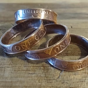 May include: Four copper rings made from US pennies are stacked on a wooden surface. The rings display the words "UNITED STATES", "ONE CENT", and "AMERICA". The rings have a worn, vintage appearance.