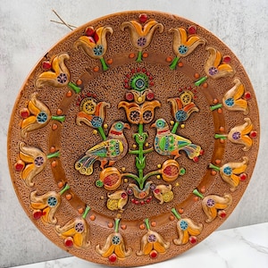 May include: A round decorative wall plate with a textured brown surface featuring a central motif of two birds perched on a stylized plant. The border is adorned with a repeating pattern of yellow tulip-like flowers with colorful accents.
