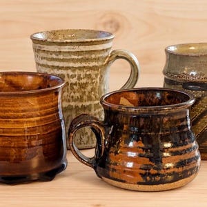 May include: Four ceramic mugs with distinct glazes. One is glossy brown, one is tall and textured with a light green wash, one is short with black and brown horizontal stripes, and one features a dark green and brown diagonal pattern.