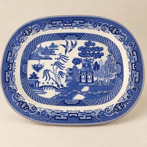 May include: An oval ceramic platter featuring a blue and white willow pattern. The design depicts a landscape with a pagoda, a bridge, a willow tree, and two birds in flight. The border is decorated with a geometric lattice and floral motifs.