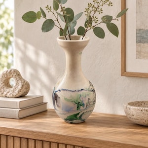 May include: A ceramic vase with a flared rim and a rounded body, featuring a cream-colored base with marbled swirls of blue, green, and black pigment. The vase contains a sprig of green eucalyptus leaves extending from the narrow neck.