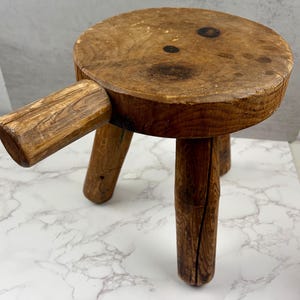 May include: A small, round, brown wooden stool featuring a thick, cylindrical handle extending from one side. The stool is supported by three sturdy, thick wooden legs and displays a natural wood grain texture with visible dark knots on the surface.