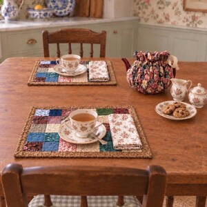May include: Two rectangular patchwork placemats featuring a grid of multicolored fabric squares, each topped with a white porcelain teacup and saucer with floral patterns, a matching folded napkin, a floral fabric tea cozy, and a small plate of cookies.
