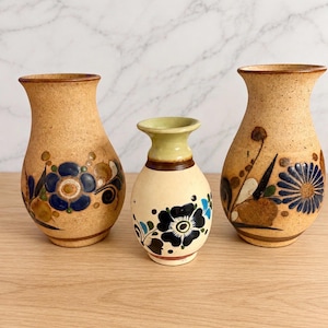 May include: Three ceramic vases of varying sizes. The two larger vases have a speckled tan finish with hand-painted blue and brown floral motifs. The smaller central vase is cream-colored with a light green rim and dark blue floral patterns.
