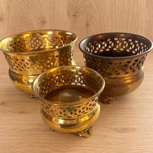 May include: Three brass bowls with a polished finish and a decorative openwork lattice pattern along the upper rim. Each bowl features a rounded base supported by three small, ornate feet, showcasing a metallic gold tone and varying levels of patina.