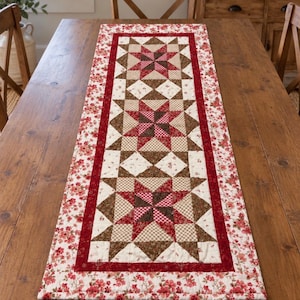 May include: A rectangular quilted table runner featuring three central star patterns in shades of red, brown, and cream. The runner is bordered by a deep red frame and an outer edge decorated with a floral print of small red flowers on a cream background.