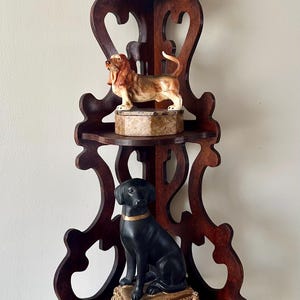 May include: A dark wood corner shelf unit with three shelves, each holding a dog figurine. The top shelf has a small white dog, the middle shelf has a brown and tan dog, and the bottom shelf has a black dog with a gold collar.