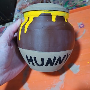 Puede incluir: Un tarro de cerámica marrón con tapa, con pintura amarilla goteando desde la parte superior. La palabra "HUNNY" está pintada en negro en la parte delantera. El tarro está sostenido por una mano sobre una superficie naranja.