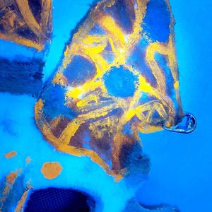 May include: Abstract art piece with a blue background and a yellow and brown design. The artwork has a unique texture and shape, with a metal clasp attached. The piece is a creative and colorful composition.