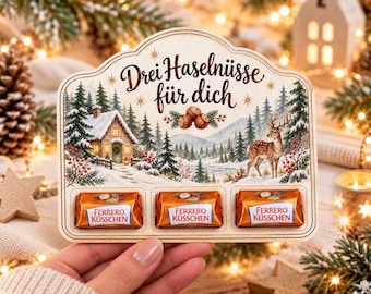 Op de afbeelding: Een decoratief houten displaybord met een winterse bosillustratie, een blokhut, een hert en de tekst "Drei Haselnüsse für dich". Het bord heeft drie rechthoekige uitsparingen met elk een Ferrero Küsschen chocoladebonbon.