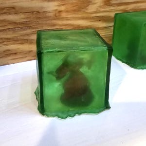 May include: Two green, cube-shaped objects. The front cube has a dark brown object encased within the transparent green material. The cubes are on a white surface with a wooden background.