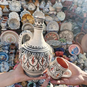 Puede incluir: Una jarra de cerámica blanca y una taza a juego con patrones geométricos negros, grises y rojos. La jarra tiene un asa y un borde decorativo. La taza tiene un asa y un interior rojo. El fondo presenta varios artículos de cerámica.