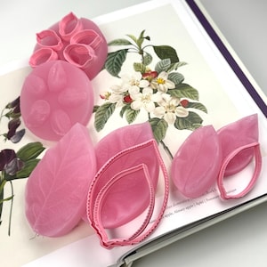 May include: A collection of pink silicone moulds and matching plastic cutters for crafting floral shapes. The set includes circular moulds with petal indentations, leaf-shaped moulds with vein textures, and corresponding serrated edge cutters.