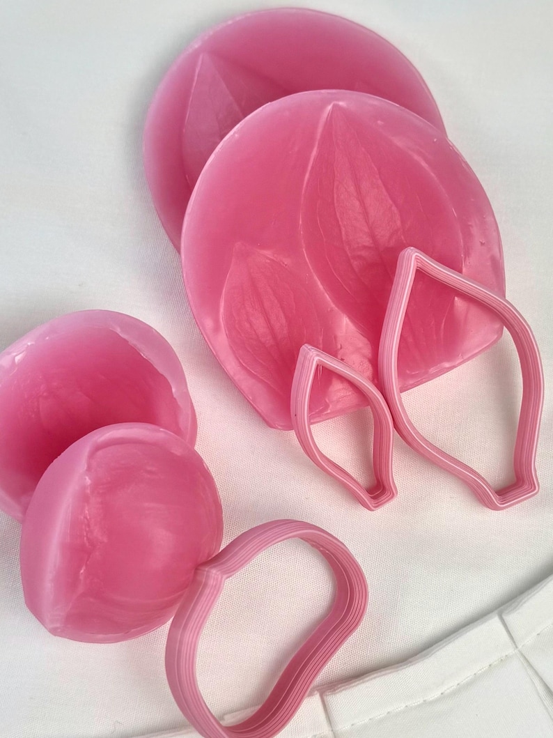 May include: A collection of pink silicone molds and matching plastic cutters for crafting. The set includes circular molds with embossed petal textures, two teardrop-shaped cutters, and one rounded cutter, all in a vibrant shade of pink.
