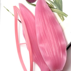 May include: A pink plastic petal mold and a matching pink plastic cutter. The mold features detailed vein textures on its surface, while the cutter has a simple, thin frame design. These tools are used for shaping floral elements in crafting.