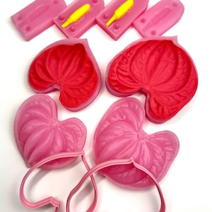 Könnte beinhalten: Eine Auswahl an rosa und roten blütenförmigen Formen und Ausstechern. Die Formen sind in verschiedenen Rosatönen gehalten, einige mit gelben Staubgefäßen. Die Ausstecher sind heller rosa, was auf ein Bastel- oder Backthema hindeutet.