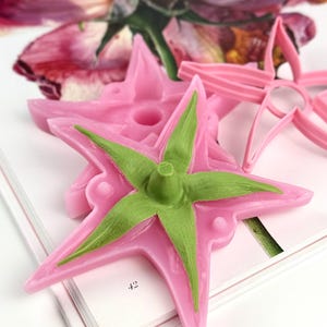 Może przedstawiać: Różowe i zielone foremki i wykrawacze w kształcie kwiatów do rękodzieła. Formy są w kolorze jasnoróżowym z zielonym środkiem, a wykrawacze w jaśniejszym różu. Przedmioty znajdują się na książce z widoczną liczbą 42.