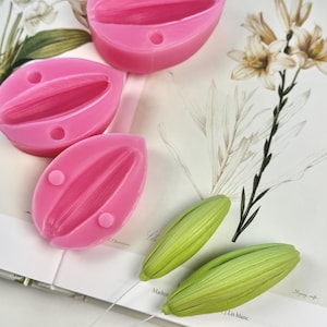 Puede incluir: Moldes de silicona rosa con forma de lirio, junto con dos réplicas de capullos de lirio verdes. Los moldes tienen interiores detallados y pequeños agujeros. Los artículos descansan sobre una página de libro con ilustraciones botánicas y texto.