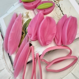 May include: A collection of pink silicone molds for crafting, including several petal-shaped molds with textured surfaces, two oval-shaped molds containing green bud-like inserts, and two thin, open-frame pink plastic cutters for shaping petals.