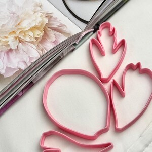 May include: A set of four pink plastic cookie cutters in various organic shapes, including one large oval and three smaller petal-like forms with pointed edges, arranged on a white surface next to an open book with a floral illustration.