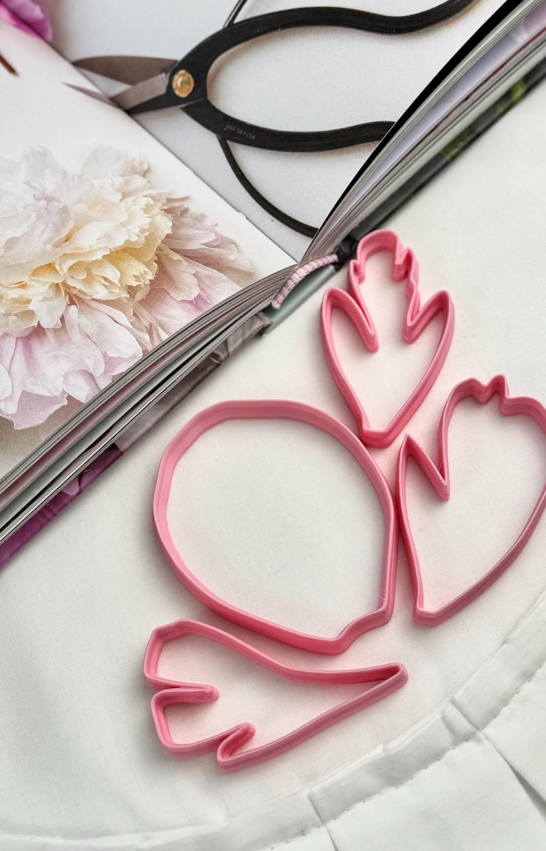 May include: A set of four pink plastic cookie cutters in various organic shapes, including one large oval and three smaller petal-like forms with pointed edges, arranged on a white surface next to an open book with a floral illustration.