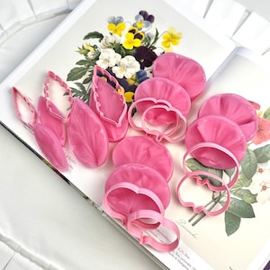 May include: A collection of pink flower petal cutters and formed petals are arranged on a floral illustration. The cutters are in various shapes, including petal and leaf designs. The petals are a vibrant pink color.