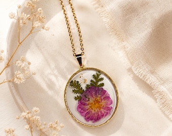May include: A gold-toned necklace featuring a circular pendant with a clear resin center. Inside the resin, a pressed purple flower and green fern leaves are embedded alongside small gold flakes. The pendant hangs from a matching gold-toned chain.
