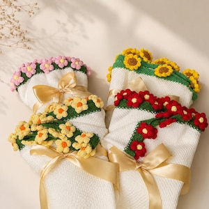 Puede incluir: Dos envoltorios de tela blanca texturizada atados con cintas de satén dorado, cada uno decorado con guirnaldas de flores de ganchillo. Uno presenta flores rosas y amarillas en tallos verdes, mientras que el otro muestra girasoles amarillos y flores rojas en tallos verdes.