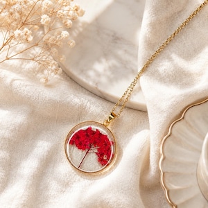 Puede incluir: Un collar con colgante circular de tono dorado que presenta una flor roja prensada incrustada en resina transparente. El colgante cuelga de una delicada cadena dorada, mostrando los pétalos rojos vibrantes sobre un fondo transparente.