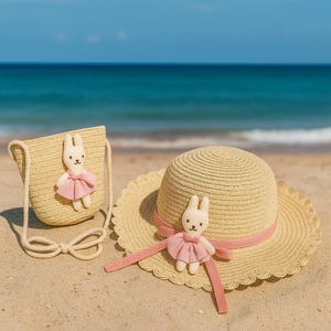 May include: A straw hat with a pink ribbon and a matching crossbody bag, both decorated with a small bunny wearing a pink dress. The items are on a sandy beach with the sea in the background.