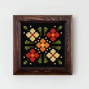 May include: A framed cross-stitch floral design. The artwork features a symmetrical pattern of flowers in shades of orange, red, and cream, set against a black background. The frame is made of dark wood.