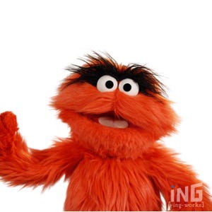 Könnte beinhalten: Eine leuchtend orangefarbene Handpuppe mit langem, zotteligem Fell, großen weißen Augen mit schwarzen Pupillen und dichten, buschigen schwarzen Augenbrauen. Die Puppe hat einen weit geöffneten Mund mit weißen Zähnen und einen erhobenen Arm.