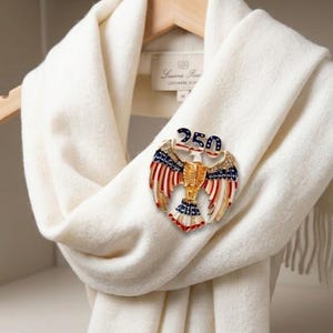 May include: A cream-colored scarf with a decorative pin. The pin is in the shape of an eagle with red, white, and blue accents, and the number "250" on top. The scarf is draped over a wooden hanger.