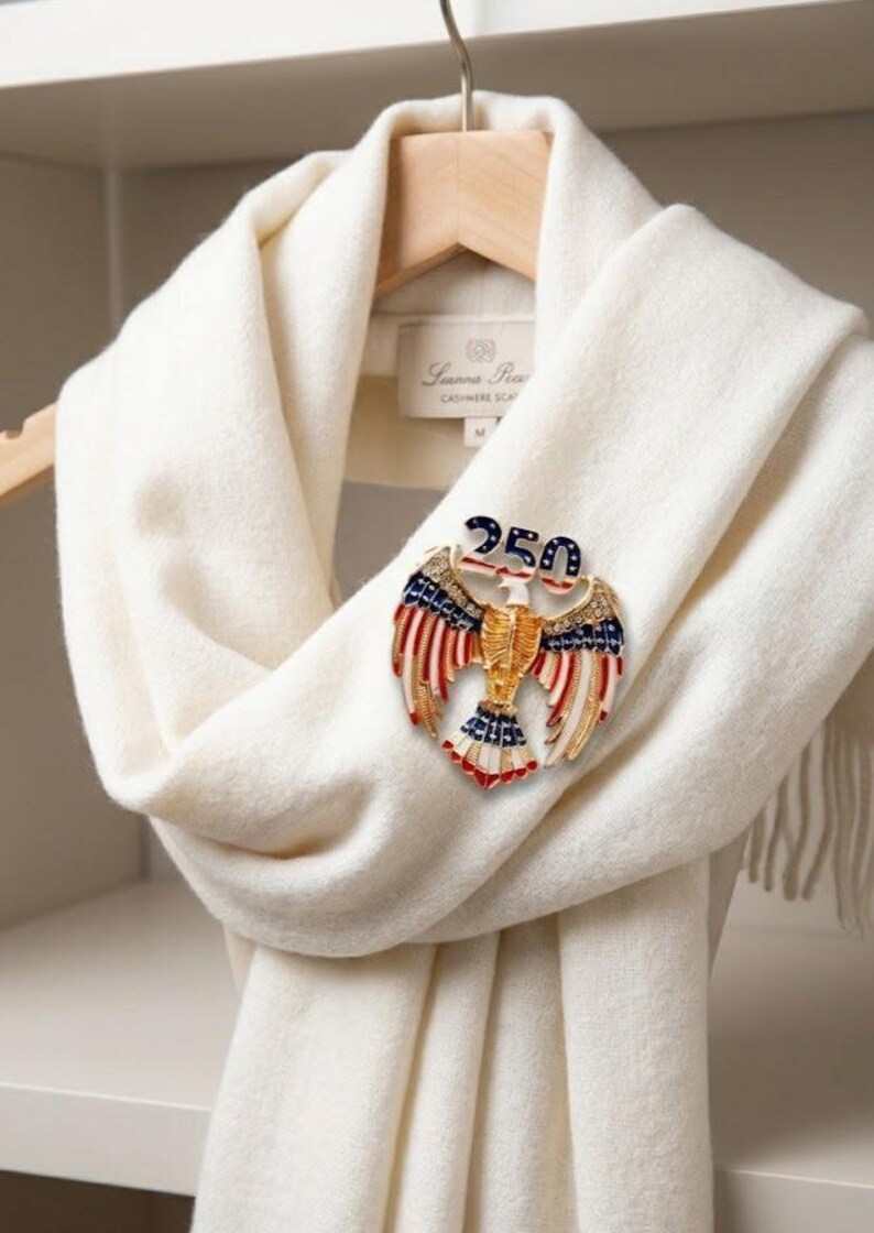 May include: A cream-colored scarf with a decorative pin. The pin is in the shape of an eagle with red, white, and blue accents, and the number "250" on top. The scarf is draped over a wooden hanger.