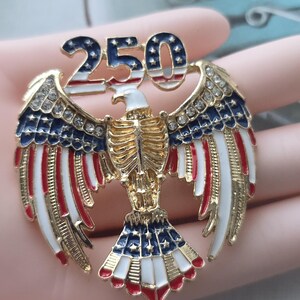 May include: A gold-toned eagle pin with outstretched wings, featuring red, white, and blue enamel details. The pin is adorned with rhinestones and the number "250" in a patriotic design, celebrating a milestone.