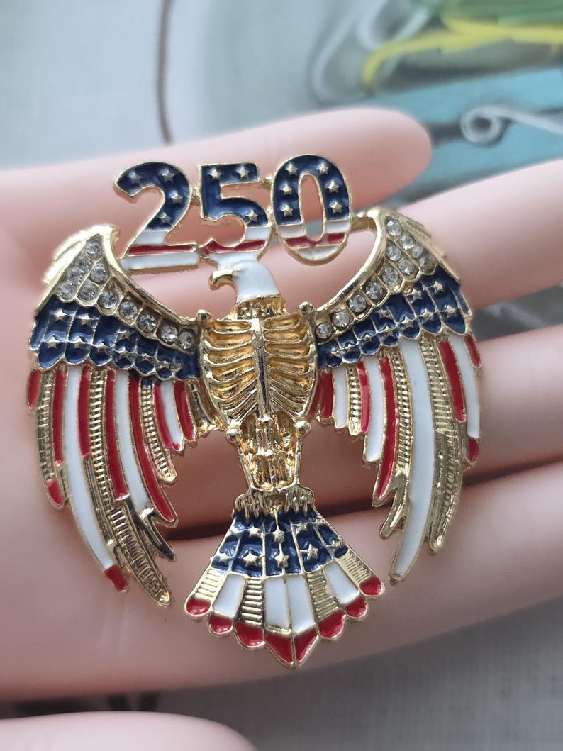 May include: A gold-toned eagle pin with outstretched wings, featuring red, white, and blue enamel details. The pin is adorned with rhinestones and the number "250" in a patriotic design, celebrating a milestone.