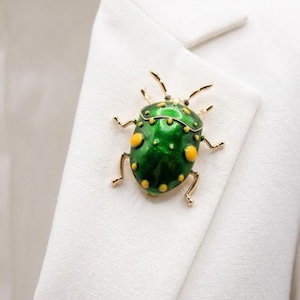 May include: A green and yellow enamel beetle brooch with gold-colored accents. The insect-shaped pin is attached to a white blazer. The brooch features a detailed design with yellow spots and delicate legs.