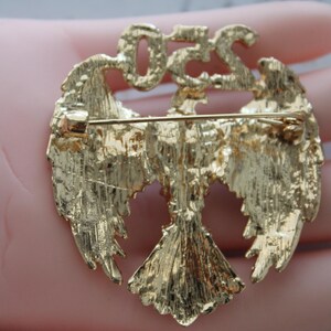 May include: Gold-toned metal brooch featuring an eagle design with outstretched wings. The brooch has the letters "OZS" at the top. The back of the brooch has a pin for attachment.