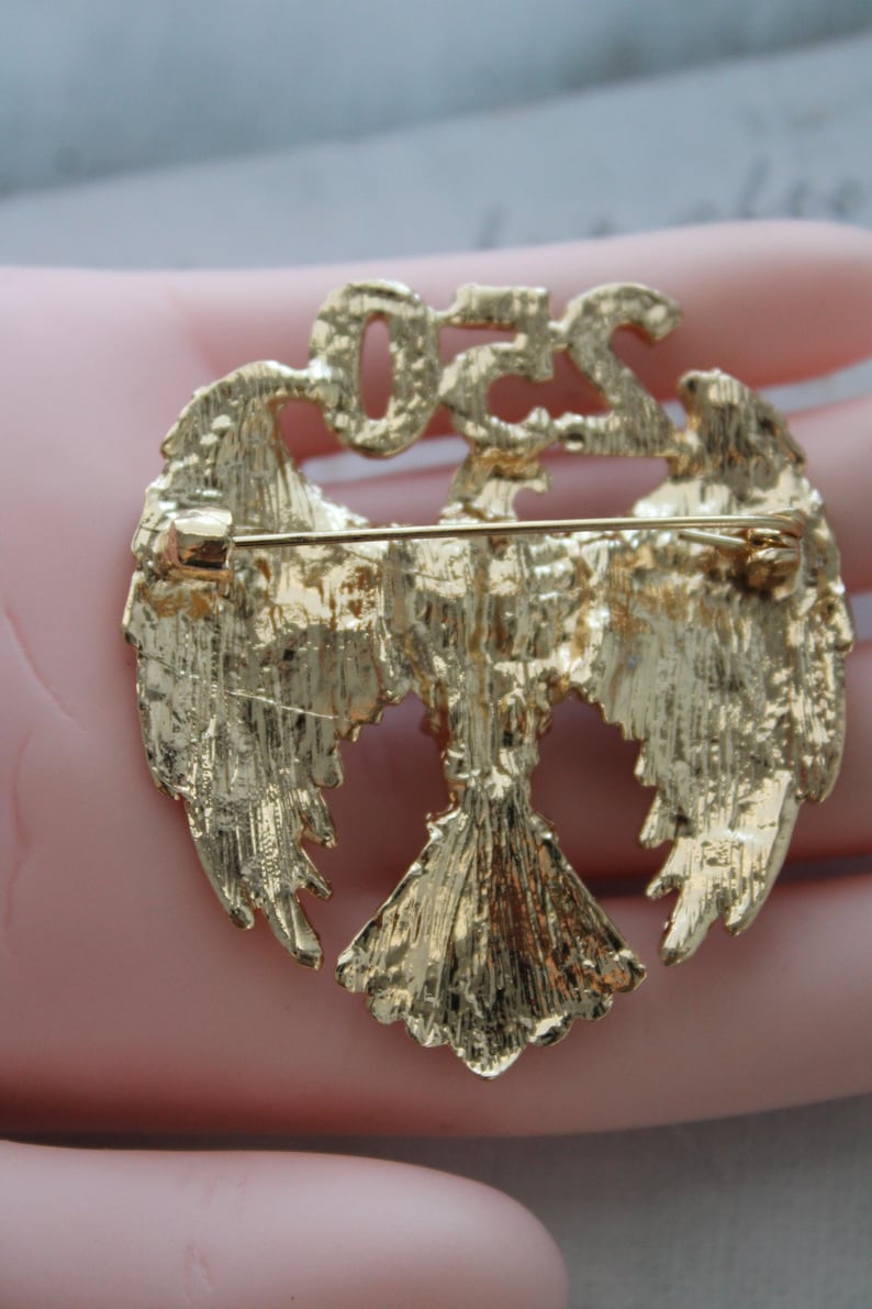 May include: Gold-toned metal brooch featuring an eagle design with outstretched wings. The brooch has the letters "OZS" at the top. The back of the brooch has a pin for attachment.