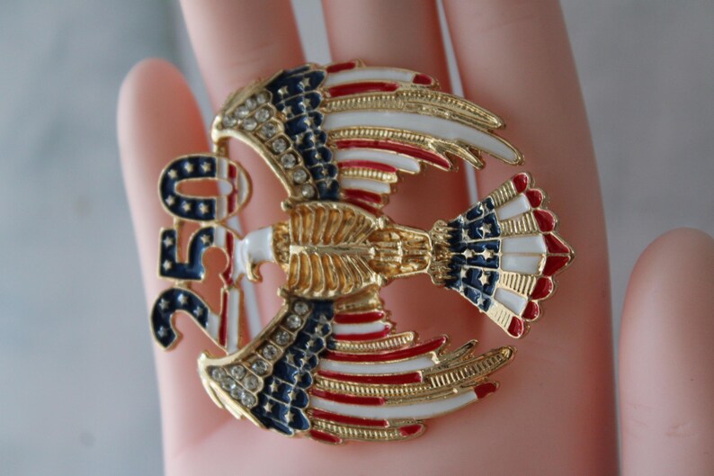 May include: A gold-toned eagle brooch with red, white, and blue enamel detailing. The brooch features the number "250" and is adorned with rhinestones. The eagle's wings and tail display the American flag design.