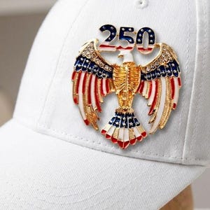 May include: White baseball cap with a patriotic eagle emblem. The emblem features a gold-colored eagle with red, white, and blue accents, and the number "250" in blue and white. The cap is displayed on a wooden stand.