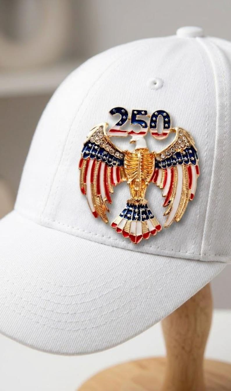 May include: White baseball cap with a patriotic eagle emblem. The emblem features a gold-colored eagle with red, white, and blue accents, and the number "250" in blue and white. The cap is displayed on a wooden stand.